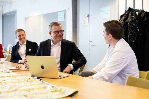 Pääministeri Petteri Orpo keskustelee pöydän ääressä lääkärin kanssa. Hänen edessään on auki oleva kannettava tietokone.