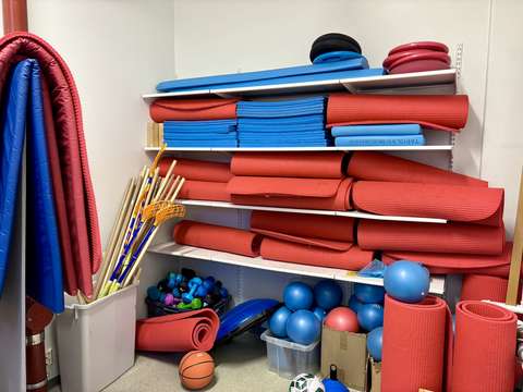 A gym storage room with four shelves holding yoga mats and exercise equipment. On the floor, there are boxes filled with various balls, exercise sticks, dumbbells, and a few floorball sticks.