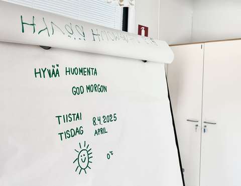 A drawing board displaying 'Good morning!' in Finnish and Swedish, along with the date and day of the week. A smiling sun is drawn on the paper.