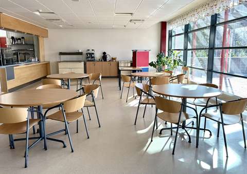 A bright and empty dining hall with large windows. Four tables with chairs are arranged in the hall, which opens up to the kitchen serving line.