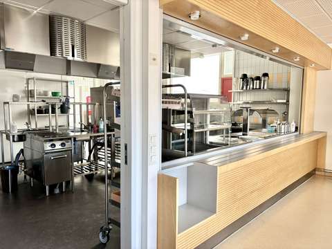 Photo taken from the dining area showing the empty cafeteria kitchen. The kitchen is equipped with stainless steel appliances and kitchen equipment.