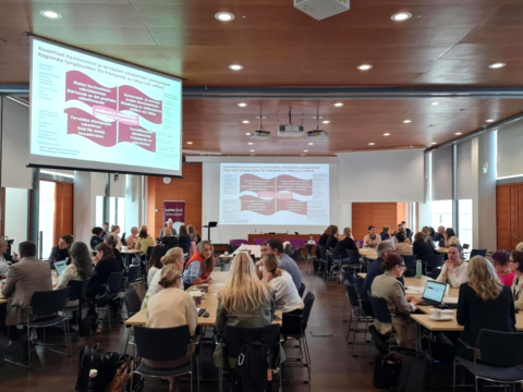 Suuri määrä ihmisiä istumassa hajautetuissa pöydissä. Heidän takanaan näkyy kalvoilla esitysdia hyvinvointia käsittelevistä teemoista.