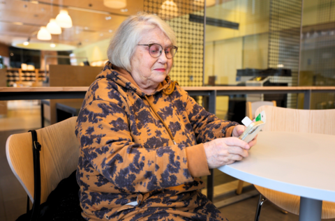 Iäkäs nainen istuu kirjastossa. Hän on pöydän ääressä ja selaa puhelinta.