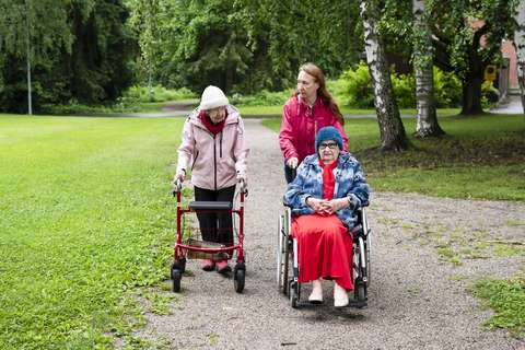 Two older women are outdoors together with a younger woman. One of the older women is seated in a wheelchair, which the younger woman is pushing. The other older woman uses a rollator to assist with mobility.
